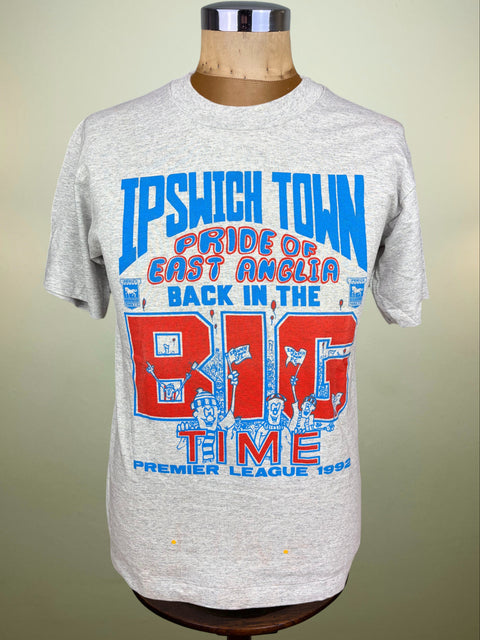 A heather gray short-sleeve t-shirt displayed on a wooden dress form mannequin. The shirt features a bold graphic print with blue and red text reading 'IPSWICH TOWN PRIDE OF EAST ANGLIA BACK IN THE BIG TIME PREMIER LEAGUE 1982'.