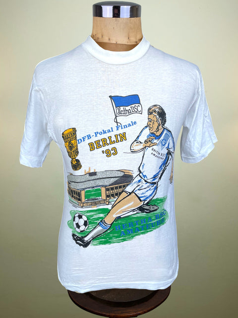 A white short-sleeve graphic T-shirt displayed on a wooden dress form mannequin. The shirt features a colorful illustration of a soccer player, a trophy, a stadium, and the text 'DFB-Pokal Finale BERLIN '93' along with a blue and white flag.