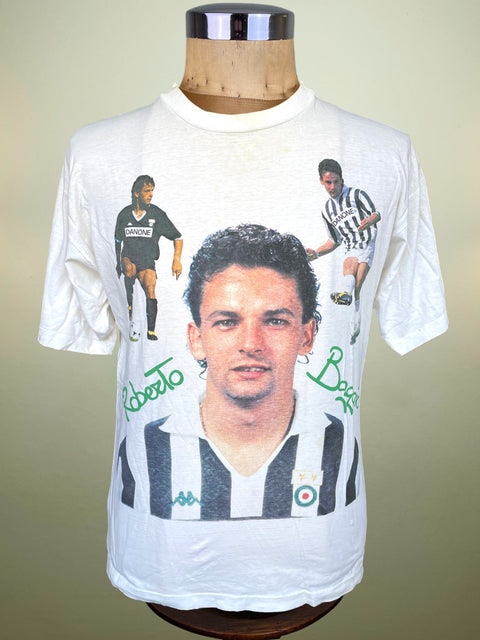 A vintage white graphic t-shirt featuring a large portrait of Roberto Baggio, with smaller action shots and green signatures, displayed on a wooden mannequin torso.