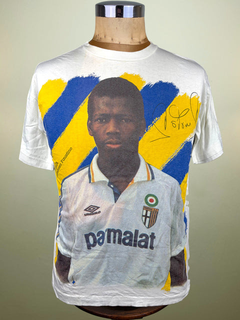 A white short-sleeve graphic T-shirt displayed on a mannequin torso against a plain light background. The shirt features a large printed portrait of a Faustino Asprilla in a white jersey, with bold blue and yellow abstract brushstroke shapes behind him and a black autograph on the upper right chest.