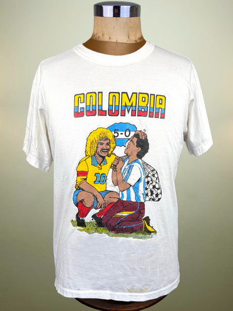 A vintage white short-sleeve t-shirt displayed on a wooden dress form mannequin. The shirt features a colorful graphic print with the word 'COLOMBIA' in rainbow letters above an illustration of two Carlos Valderrama laughing at Diego Maradona with a '5-0' score graphic.