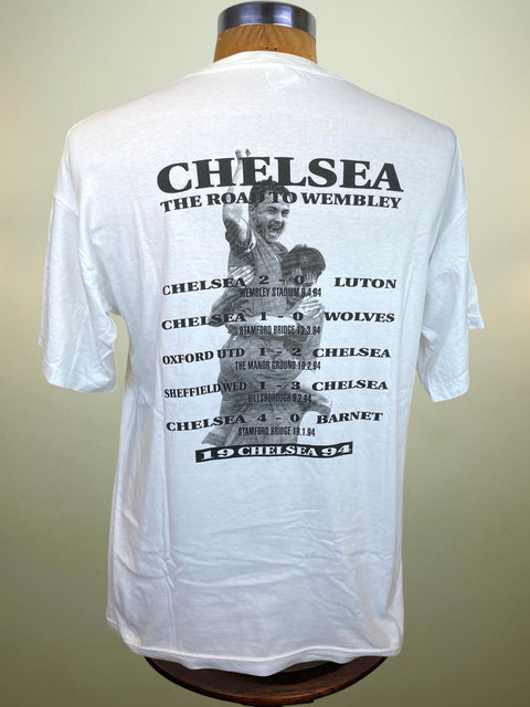 White short-sleeve T-shirt displayed on a mannequin torso, showing the back side with a large black printed football match graphic and text for Chelsea.