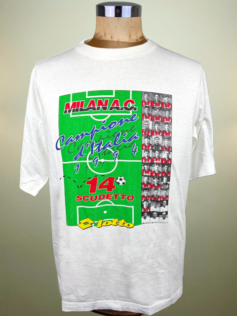 A white short-sleeve Lotto graphic t-shirt displayed on a wooden dress form mannequin. The shirt features a large green soccer field print on the front with 'MILAN 14th Scudetto'.
