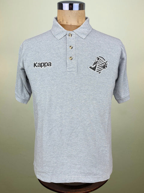 A light gray heathered Kappa polo shirt displayed on a wooden dress form mannequin against a soft beige background. The shirt features the black Kappa logo embroidered on the right chest and the club crest on the left chest.