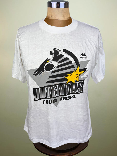 A white short-sleeve t-shirt displayed on a wooden mannequin torso. The shirt features a graphic print for the Juventus football club's 'Tour 1994' with a zebra head, black stripes, a yellow star, and the Kappa logo.