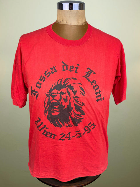 A red short-sleeve t-shirt displayed on a wooden mannequin torso against a light beige background. The shirt features a large black graphic print of a lion's head with the text 'Fossa dei Leoni' and 'Wien 24.