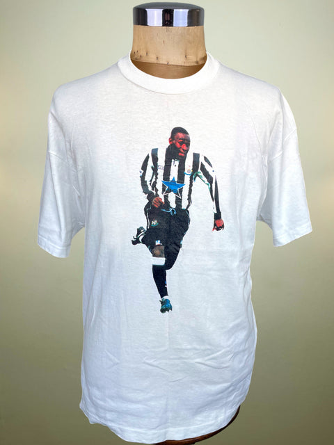 A white short-sleeve t-shirt displayed on a wooden dress form mannequin. The shirt features a graphic print of soccer player Andy Cole wearing a black and white striped Newcastle United jersey with a blue star on the chest.