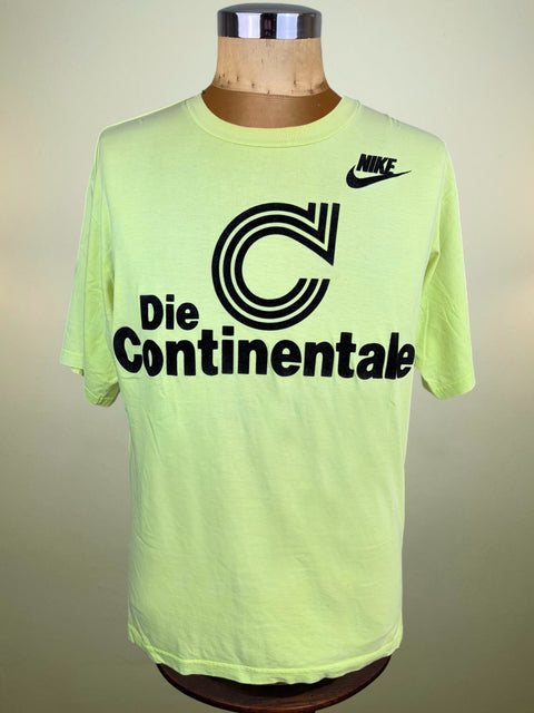A neon yellow-green Nike t-shirt displayed on a wooden dress form mannequin. The shirt features a large black graphic print on the front reading 'Die Continentale' with a stylized 'C' logo, and a black Nike swoosh on the upper right chest.