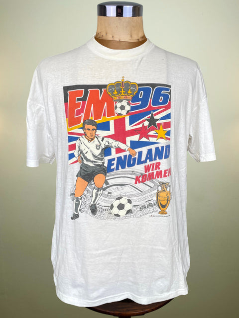 A white vintage Germany 1996 soccer t-shirt displayed on a wooden dress form mannequin. The front features a colorful graphic with the text 'EM 96 ENGLAND WIR KOMMEN', a soccer player illustration, the Union Jack flag, and a gold trophy.