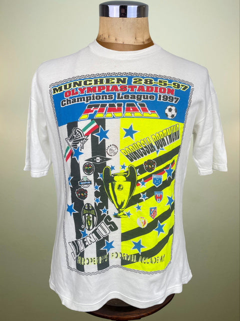 A white short-sleeve graphic T-shirt displayed on a wooden mannequin torso against a plain light background. The shirt features a large front print celebrating the 1997 Champions League final in Munich, with black-and-white stripes, a bright yellow panel, blue stars, and the text 'Munich 28-5-97 Olympiastadion Champions League 1997 Final.