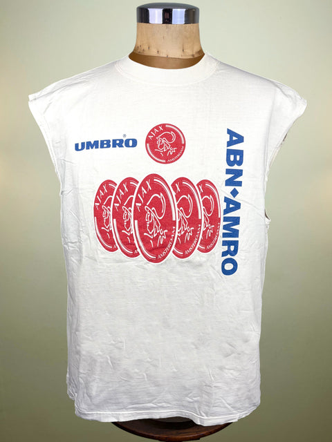 A white sleeveless t-shirt displayed on a wooden mannequin torso. The shirt features blue 'UMBRO' text, a red circular logo, a row of red circular graphics with white signatures, and vertical blue 'ABN-AMRO' text.