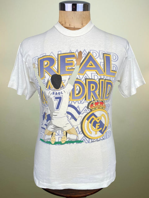 A white Real Madrid graphic t-shirt featuring Raul illustration and team crest, displayed on a wooden mannequin torso against a neutral beige background.