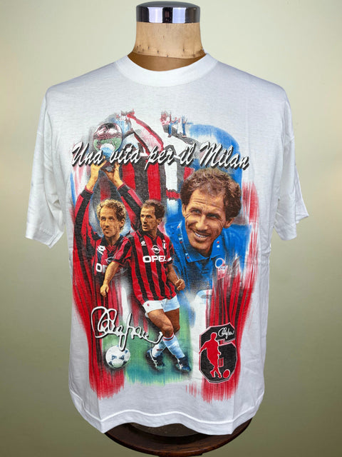 A white short-sleeve graphic T-shirt displayed on a mannequin torso against a plain beige background. The shirt features a large multicolor print with portraits of Franco Baresi, red and black striped jerseys, blue tones, and the text 'Una vita per il Milan' plus the AC Milan crest.