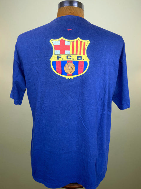 A blue short-sleeve t-shirt displayed on a wooden mannequin torso, showing the back with a large FC Barcelona crest and a small Nike swoosh above it.