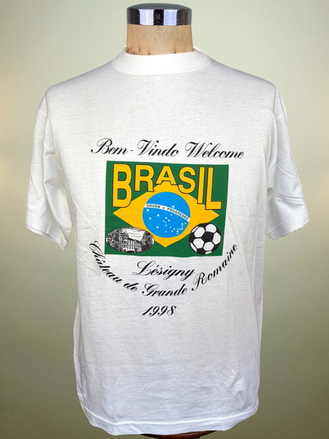 A white short-sleeve t-shirt displayed on a wooden mannequin torso against a light beige background. The shirt features a colorful graphic on the front with the text 'Bien-Vindo Welcome BRASIL' and 'Chateau de Grande Roncane 1998' surrounding a green, yellow, and blue Brazil flag design.