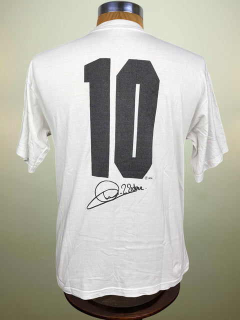 A white short-sleeve t-shirt displayed on a wooden mannequin torso, showing the back with a large black number 10 and Zinedine Zidane's signature graphic.