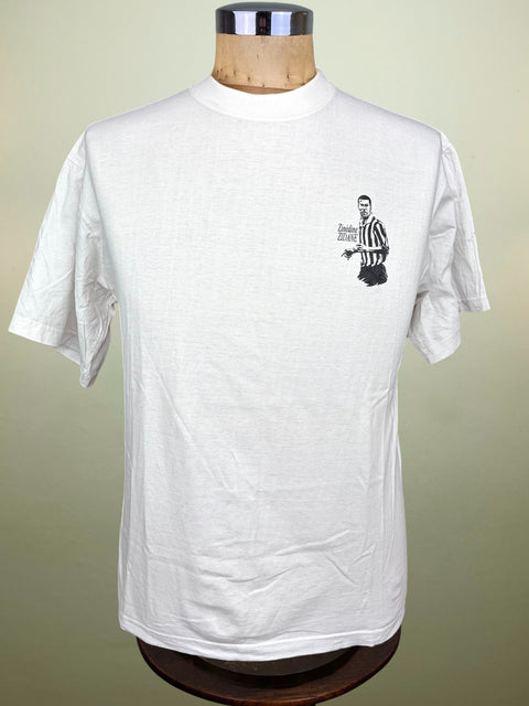 A white short-sleeve crewneck t-shirt displayed on a wooden mannequin torso. The shirt features a small black graphic on the left chest depicting Zinedine Zidane