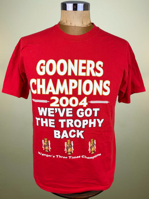 A red short-sleeve graphic T-shirt displayed on a wooden mannequin torso against a plain light beige background. The shirt features large white text reading 'GOONERS CHAMPIONS 2004 WE'VE GOT THE TROPHY BACK' with small illustrated trophy graphics near the bottom.