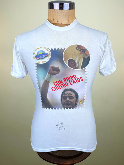 A white short-sleeve graphic T-shirt displayed on a wooden dress form mannequin against a beige studio background. The shirt features a large front print with a postage-stamp border, a circular inset photo, a raised fist, the text 'CON PIPPO CONTRO L'AIDS' in red, and a small circular logo near the left chest.